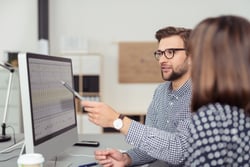 Proficient young male employee with eyeglasses and checkered shirt, explaining a business analysis displayed on the monitor of a desktop PC to his female colleague, in the interior of a modern office-2